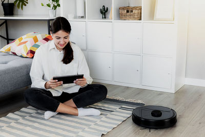 Young caucasian woman using tablet, chatting, playing while robot vacuum cleaner is cleaning floor