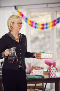 Senior woman holding gift box and juice while standing by table at home