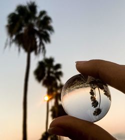 Close-up of hand holding crystal ball against sky during sunset