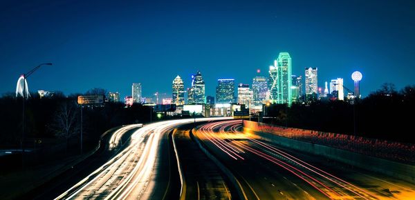 Light trails on road amidst buildings against sky at night