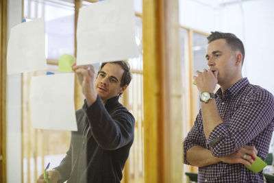 Male colleagues sticking notes on glass in office