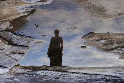 Reflection of woman in puddle standing on rock against sky