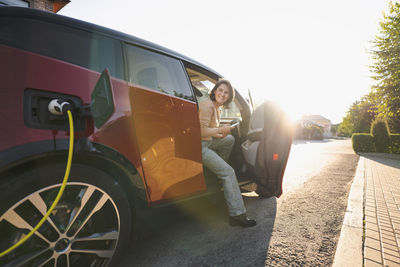 Happy woman with mobile phone sitting in electric car at vehicle charging station on sunny day