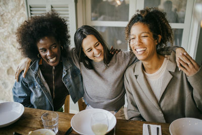 Cheerful woman sitting with arm around female friends at dining table enjoying dinner in patio