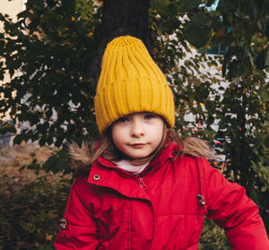 Portrait of a smiling girl in a red jacket and yellow hat against the backdrop of foliage