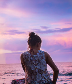 Rear view of young woman looking at sea while sitting on beach against sky during sunset