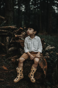 Cute boy looking away while sitting on wood in forest