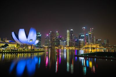 Illuminated modern buildings by river against sky at night