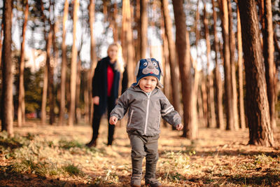 Baby boy walking in park in autumn with his mother standing behind him
