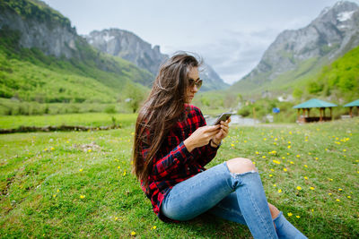 Woman using phone while sitting on grassy field against mountains