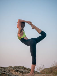 Side view of woman doing exercise on rock against sky during sunset