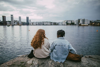 Rear view of male and female teenage friends sitting on rock near sea in city