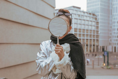 Young woman holding mirror while standing against buildings in city