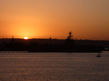 Silhouette cranes by sea against orange sky