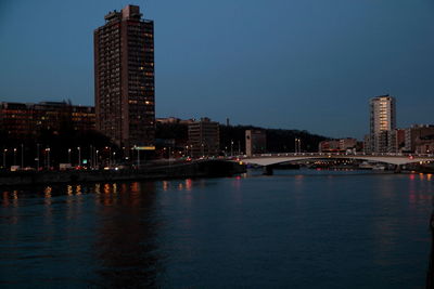 Illuminated buildings by river against sky at dusk