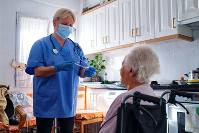 Caring nurse in uniform and protective mask with syringe in hands standing near elderly disabled woman in wheelchair in kitchen