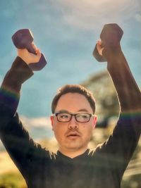 Low angle view of young asian man lifting dumbbells above the head against blue sky and trees