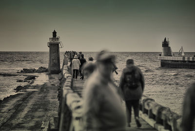 People walking on footbridge over sea by lighthouse against clear sky