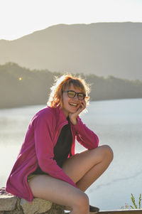 Portrait of smiling woman sitting by water against sky