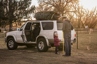 Daughter looking at father while standing against car with christmas tree on roof