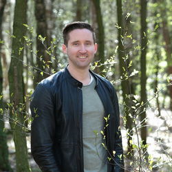 A young men with blue jeans, black jacket and white shoes in the forest.