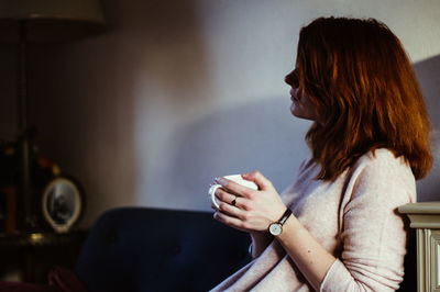 Side view of woman holding coffee cup while sitting on sofa