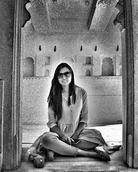 Portrait of young woman wearing sunglasses sitting against wall