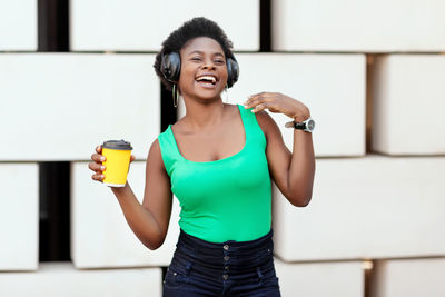 Charming african woman holds coffee in a disposable cup, listens to a pleasant melody in headphones 