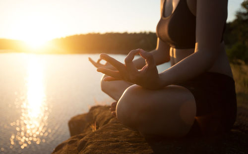 Low section of woman meditating while sitting on rock at lakeshore during sunset