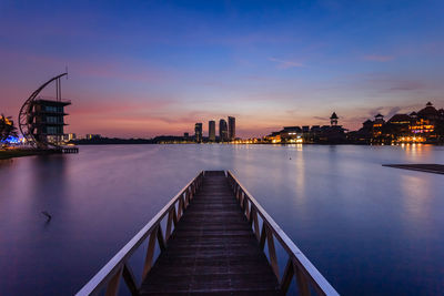 Bridge over river by illuminated buildings against sky at sunset