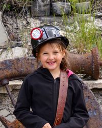 Portrait of smiling girl wearing hardhat and belt while standing against rusty metal