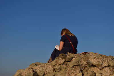 Rear view of woman sitting on rock against clear blue sky