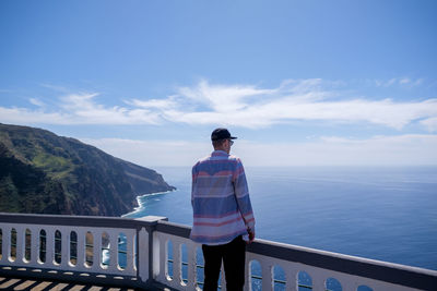 Rear view of man standing on railing by sea against sky