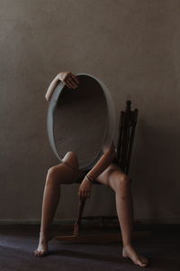 Low section of naked woman holding mirror while sitting on chair in room