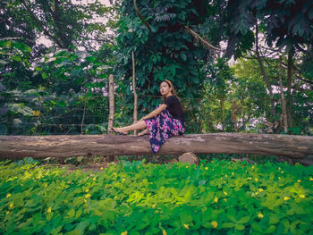 Young woman sitting on log amidst trees and plants