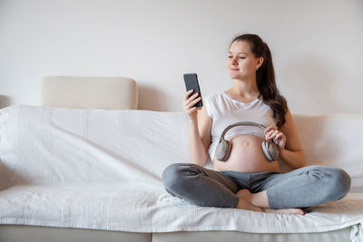 Young pregnant woman holding headphones on stomach while sitting on sofa at home