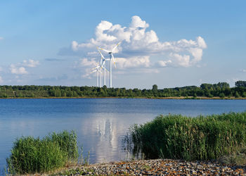 Several wind turbines stand in front of a huge cloud formation at the shore of a lake