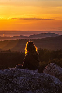 Rear view of teenage girl sitting on rock against sky during sunset