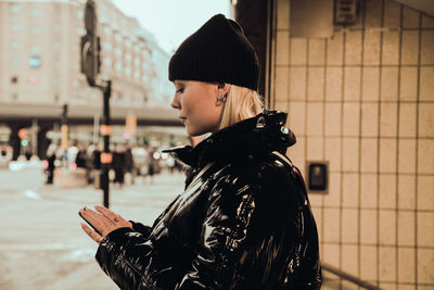 Side view of young woman using mobile phone while wearing black shiny jacket at subway's entrance in city during winter