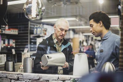 Male customer with kettle standing by salesman seen through glass window