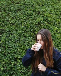 Young woman with long hair drinking coffee against bush at park