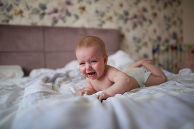 Charming baby, 6 months old, lies crying on his stomach on a bed in a bright, real interior,