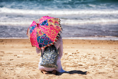 Rear view of woman with umbrella kneeling at beach during sunny day