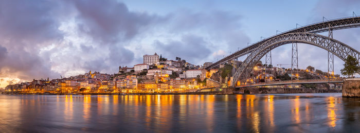 Illuminated bridge over river by buildings against sky