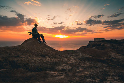 Full length of man holding skateboard sitting against sky during sunset