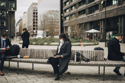 Female entrepreneur working through laptop while sitting with colleagues on bench during pandemic