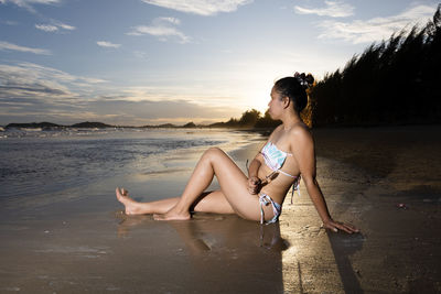 Side view of woman sitting on beach against sky during sunset