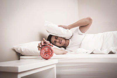 Young man pressing head with pillow while closing alarm clock