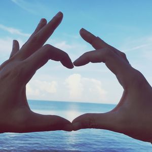 Cropped hands making heart shape by sea against sky