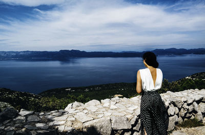 Rear view of woman sitting on rock by sea against sky